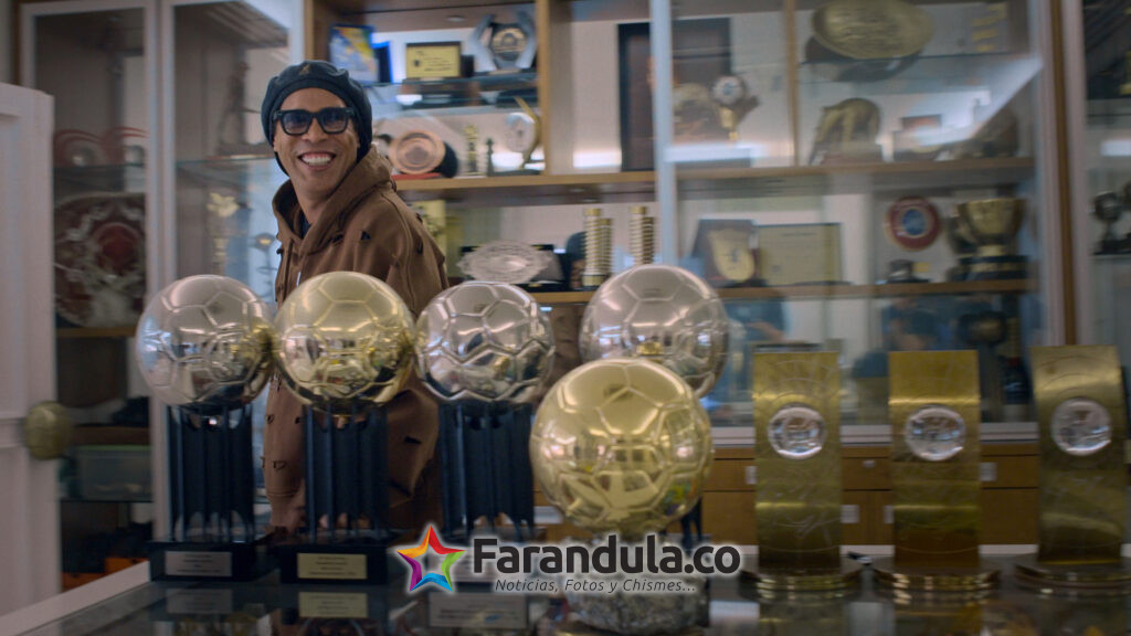 "Ronaldinho Gaúcho smiling and wearing glasses stands in a trophy room filled with numerous awards and medals displayed in glass cases and on shelves, with several golden soccer trophies prominently on a table in the foreground."